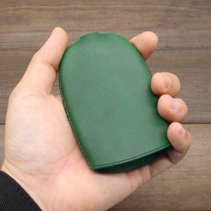 May include: A dark green leather key case held in a hand. The case is oval-shaped with a stitched border. The background is a wooden surface.