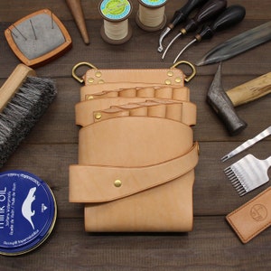 Puede incluir: Un cintur&oacute;n de herramientas de cuero de color beige claro con m&uacute;ltiples bolsillos y una correa, con herrajes de lat&oacute;n. Varias herramientas de marroquiner&iacute;a, incluyendo martillos, cepillos y agujas, est&aacute;n dispuestas alrededor del cintur&oacute;n sobre una superficie de madera. Tambi&eacute;n hay una lata de aceite de vis&oacute;n.