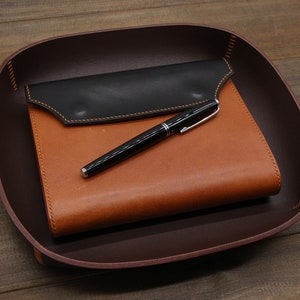 May include: A brown leather tray with a black leather notebook and a silver pen inside.