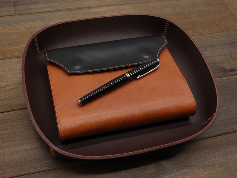 May include: A brown leather tray with a black leather notebook and a silver pen inside.