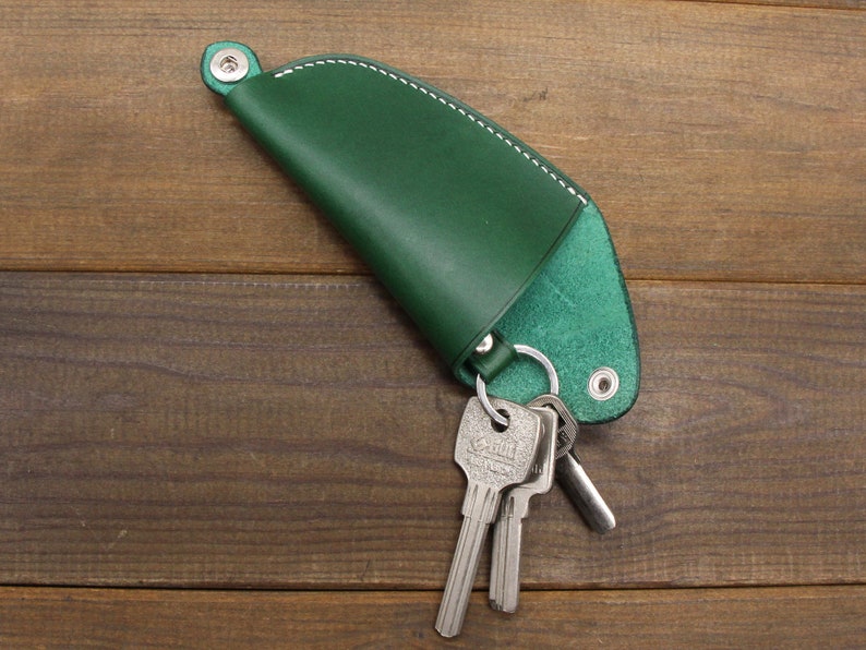 May include: A green leather key holder with a white stitch detail and a silver key ring. The key holder is shaped like a crescent moon and has a flap that folds over to secure the keys.