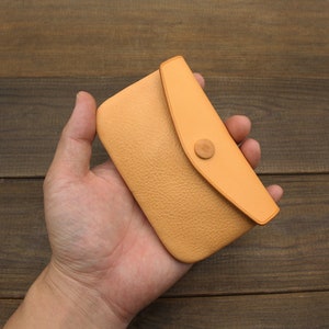 Puede incluir: Una cartera de monedas de cuero marr&oacute;n claro con cierre de solapa y bot&oacute;n de madera. La cartera se sostiene en una mano.