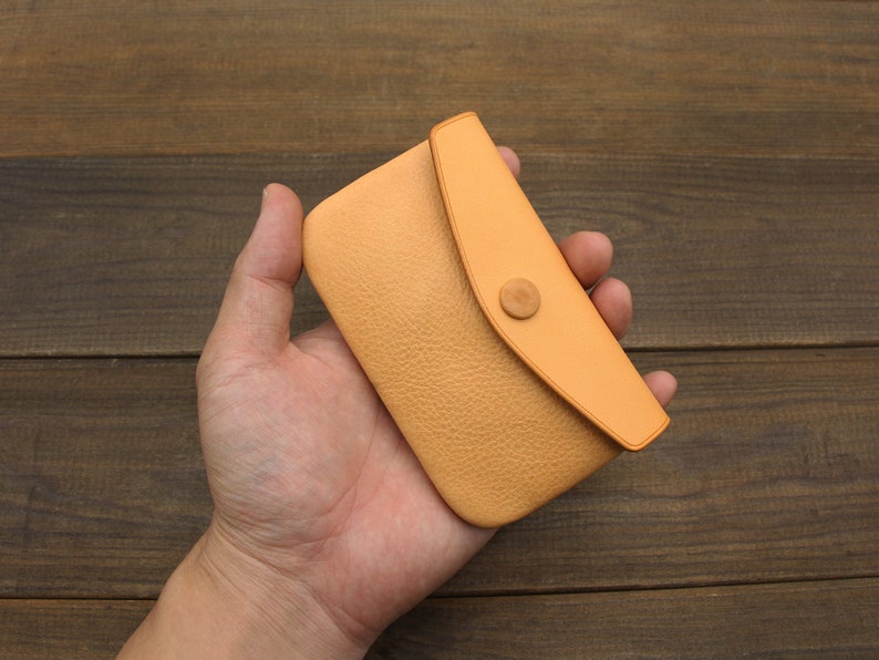 Puede incluir: Una cartera de monedas de cuero marr&oacute;n claro con cierre de solapa y bot&oacute;n de madera. La cartera se sostiene en una mano.