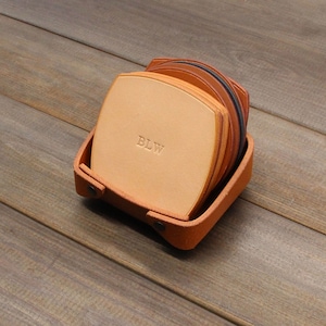 Puede incluir: Un juego de seis posavasos de cuero en un soporte de cuero marr&oacute;n. Los posavasos est&aacute;n apilados y el posavasos superior tiene las iniciales "BLW" grabadas.