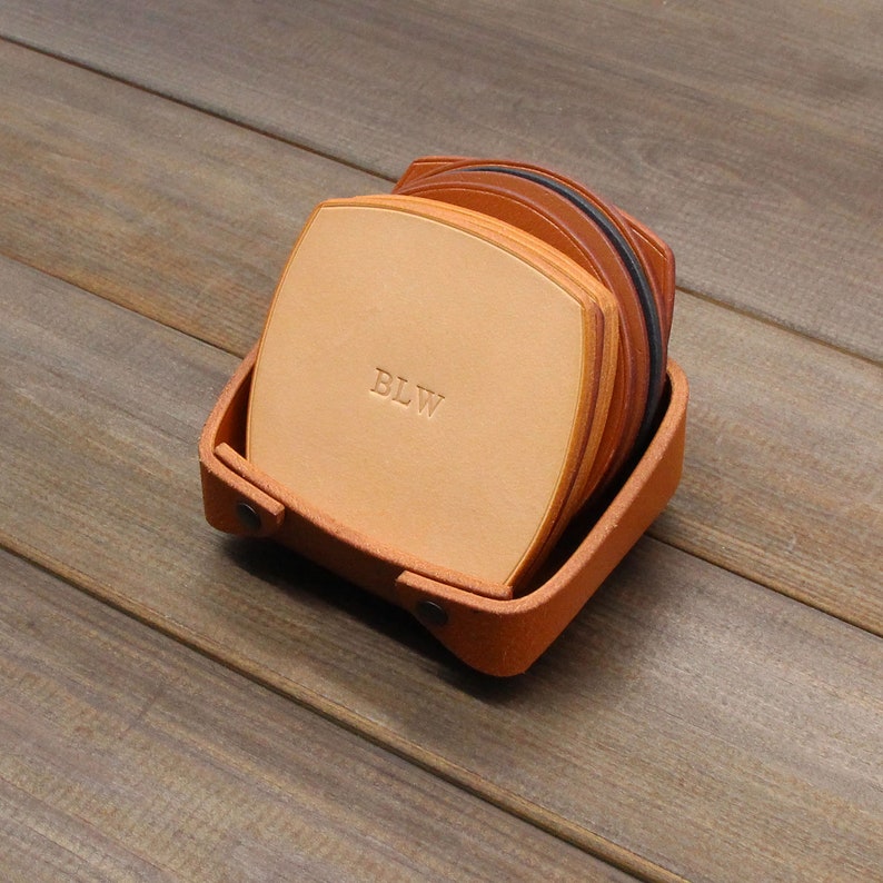 Puede incluir: Un juego de seis posavasos de cuero en un soporte de cuero marr&oacute;n. Los posavasos est&aacute;n apilados y el posavasos superior tiene las iniciales "BLW" grabadas.