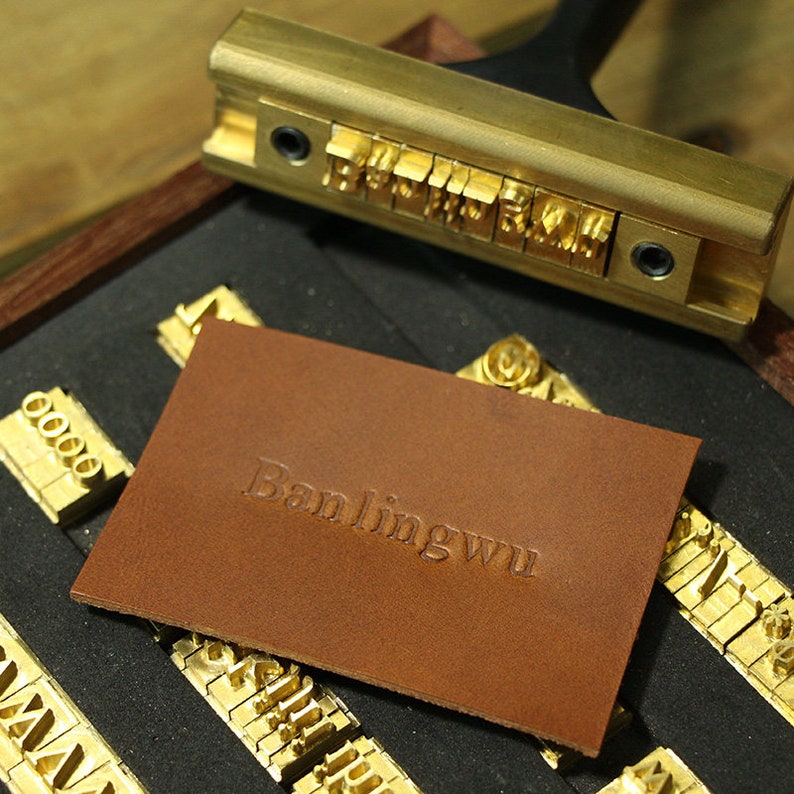 Puede incluir: Un parche de cuero marr&oacute;n con el texto "Banlingwu" estampado en dorado. El parche est&aacute; rodeado de una variedad de sellos de letras met&aacute;licas en una caja de madera.