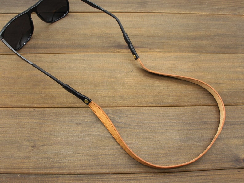 May include: Sunglasses with black frames and dark lenses are attached to a tan leather strap. The strap has black leather connectors with gold-colored rivets. The background is a wooden surface.