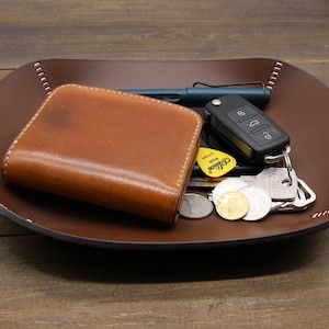 May include: A brown leather tray with a stitched edge, holding a brown leather wallet, a set of keys with a key fob, a pen, and several coins.