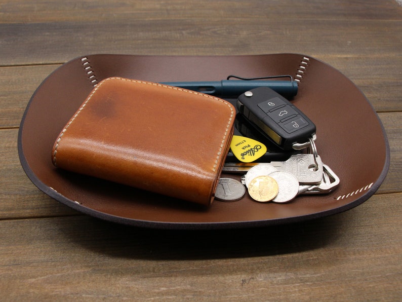 May include: A brown leather tray with a stitched edge, holding a brown leather wallet, a set of keys with a key fob, a pen, and several coins.