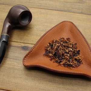 May include: A dark brown wooden tobacco pipe with a black stem sits next to a triangular, brown leather tray filled with loose tobacco. The items are on a wooden surface.