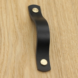 May include: Black leather cabinet pull with two brass screws. The pull is curved and has a simple design.
