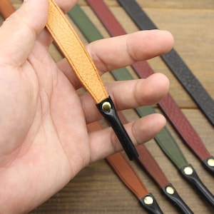 May include: Close-up of several leather wrist straps in various colors, including tan, brown, green, burgundy, and black. Each strap has a black end piece and a gold-colored rivet. The straps are displayed against a wooden background.