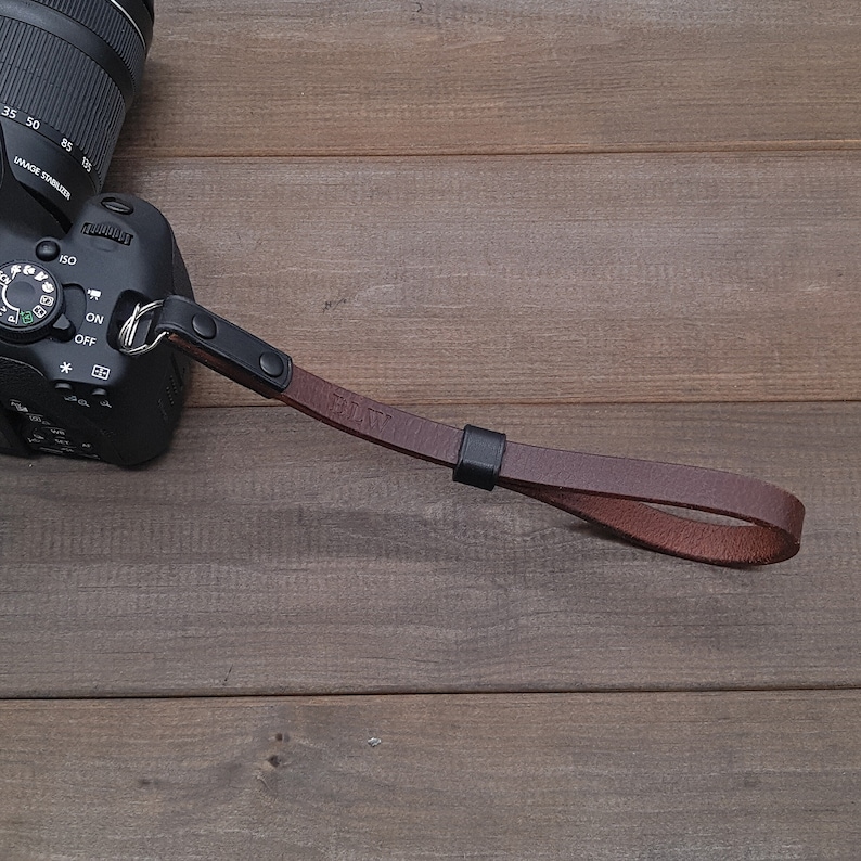 May include: A brown leather camera strap with black metal hardware. The strap has the initials "BLW" embossed on it.