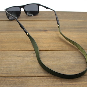 May include: Black sunglasses with a dark green leather strap. The strap is attached to the sunglasses' arms with small gold-colored rivets. The sunglasses are resting on a wooden surface.
