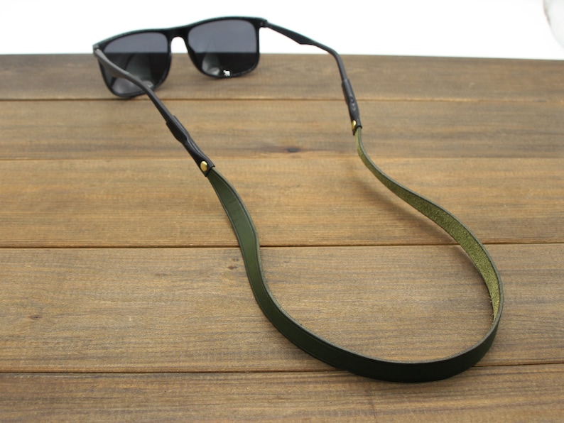 May include: Black sunglasses with a dark green leather strap. The strap is attached to the sunglasses' arms with small gold-colored rivets. The sunglasses are resting on a wooden surface.