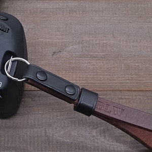 Pu&ograve; includere: Un cinturino per fotocamera in pelle marrone con dettagli neri e le iniziali "BLW" in rilievo sul cinturino. Il cinturino &egrave; attaccato a una fotocamera nera.