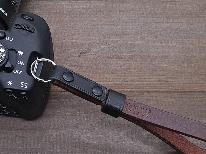 Pu&ograve; includere: Un cinturino per fotocamera in pelle marrone con dettagli neri e le iniziali "BLW" in rilievo sul cinturino. Il cinturino &egrave; attaccato a una fotocamera nera.