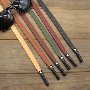 May include: Black sunglasses rest on a wooden surface, accompanied by several leather eyeglass straps in various colors: tan, brown, dark brown, green, burgundy, and black. Each strap has a black rubber end and a gold-colored metal accent.