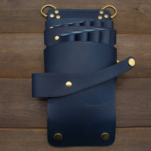 Puede incluir: Un organizador de herramientas de peluquer&iacute;a de cuero azul marino con remaches y anillos dorados. El organizador tiene m&uacute;ltiples compartimentos y un cierre de correa. El texto "Foushee 97" est&aacute; grabado en la parte delantera.