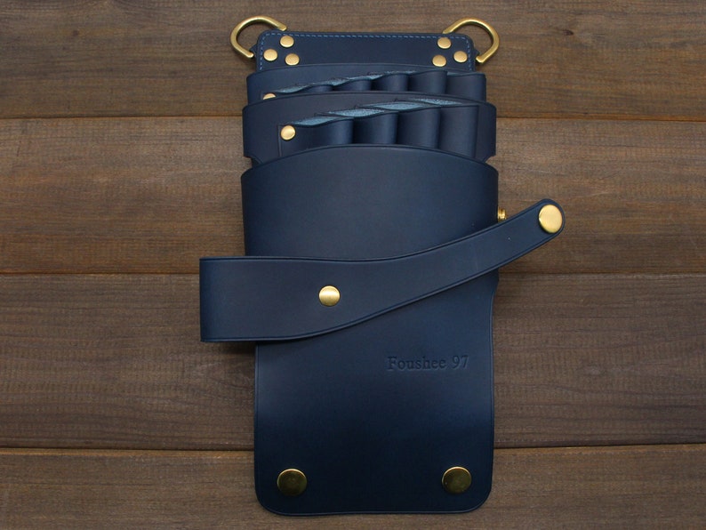 Puede incluir: Un organizador de herramientas de peluquer&iacute;a de cuero azul marino con remaches y anillos dorados. El organizador tiene m&uacute;ltiples compartimentos y un cierre de correa. El texto "Foushee 97" est&aacute; grabado en la parte delantera.