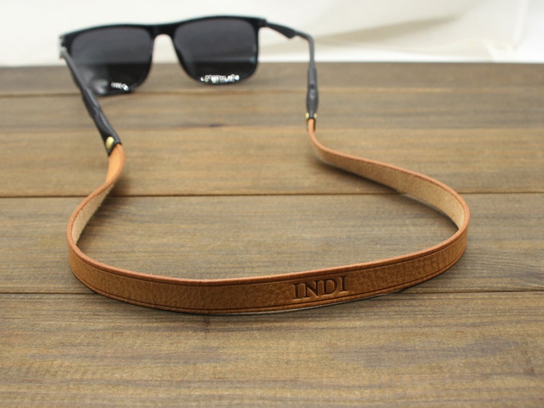 May include: Sunglasses with black frames and a brown leather strap. The strap is embossed with the name "INDI". The strap is designed to hold the sunglasses around the neck. The strap is made of a light brown leather.