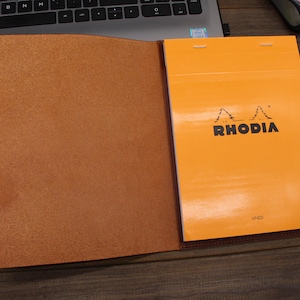 May include: A brown leather notebook cover with a bright orange lined notebook inside. The notebook cover has a flap that closes with a snap. The notebook has the word "RHODIA" printed on the front in black.