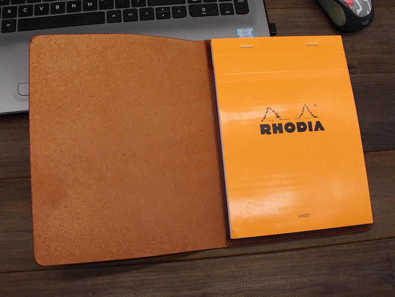 May include: A brown leather notebook cover with a bright orange lined notebook inside. The notebook cover has a flap that closes with a snap. The notebook has the word "RHODIA" printed on the front in black.