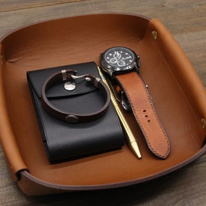 May include: A brown leather tray with a black leather wallet, a silver bracelet, a watch with a black face and brown band, and a gold pen.