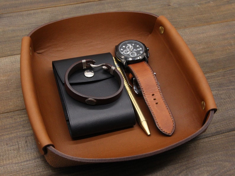 May include: A brown leather tray with a black leather wallet, a silver bracelet, a watch with a black face and brown band, and a gold pen.
