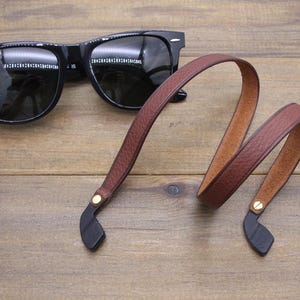 May include: Black sunglasses with a brown leather strap. The strap has black ends and gold-colored rivets. The sunglasses are on a wooden surface.