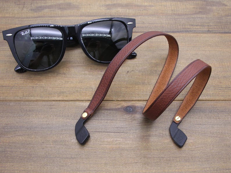 May include: Black sunglasses with a brown leather strap. The strap has black ends and gold-colored rivets. The sunglasses are on a wooden surface.
