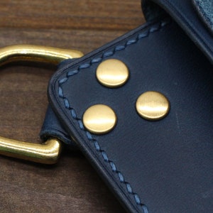 Puede incluir: Primer plano de una cartera de cuero azul marino con herrajes met&aacute;licos dorados. La cartera presenta tres remaches dorados y una anilla en D grande. La costura es de color azul claro contrastante.