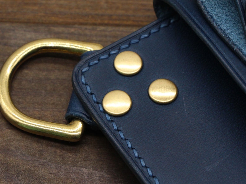 Puede incluir: Primer plano de una cartera de cuero azul marino con herrajes met&aacute;licos dorados. La cartera presenta tres remaches dorados y una anilla en D grande. La costura es de color azul claro contrastante.