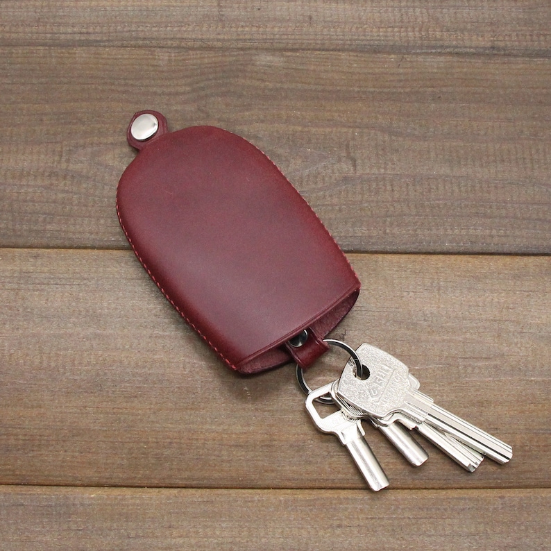 May include: A burgundy leather key case with a silver snap closure and a metal key ring holding several silver keys. The key case is oval-shaped and sits on a wooden surface.