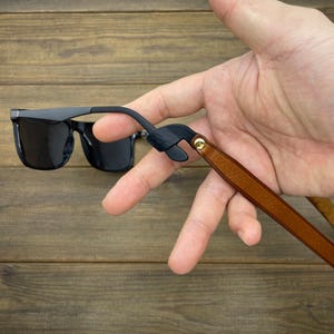 May include: Black sunglasses with a dark grey frame and a brown leather strap. The strap is attached to the sunglasses with a black rubber piece and a gold-colored rivet. The sunglasses are held against a wooden background.