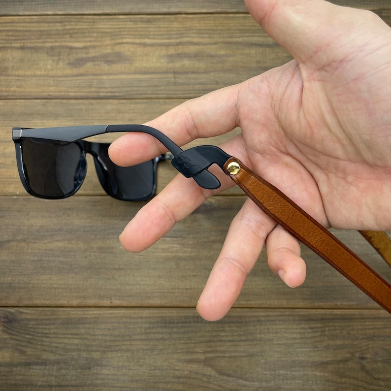 May include: Black sunglasses with a dark grey frame and a brown leather strap. The strap is attached to the sunglasses with a black rubber piece and a gold-colored rivet. The sunglasses are held against a wooden background.