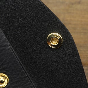 Puede incluir: Primer plano de un bolso de cuero negro con un cierre de presi&oacute;n de metal dorado. El broche tiene el texto "TRF" grabado.