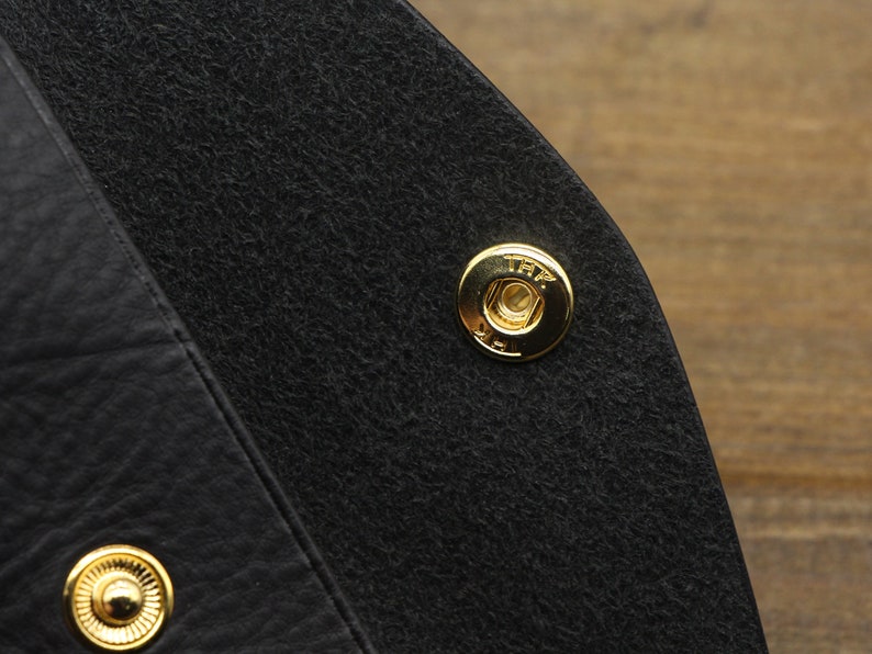 Puede incluir: Primer plano de un bolso de cuero negro con un cierre de presi&oacute;n de metal dorado. El broche tiene el texto "TRF" grabado.