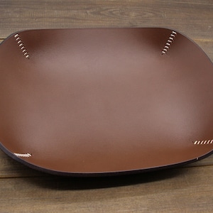 May include: A brown leather tray with white stitching around the edges. The tray is square shaped and has a slightly rounded bottom.