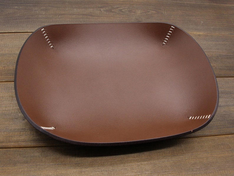 May include: A brown leather tray with white stitching around the edges. The tray is square shaped and has a slightly rounded bottom.