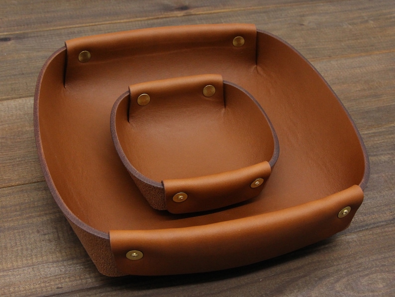 May include: Two brown leather trays with brass rivets. The smaller tray is nested inside the larger tray.