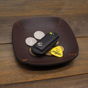 Puede incluir: Una bandeja de cuero marr&oacute;n con costuras naranjas. La bandeja contiene un encendedor negro, tres monedas de plata y una p&uacute;a de guitarra amarilla con el texto "Guitar Pick 0.71mm" y la palabra "D'Addario".