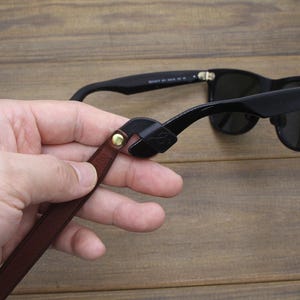 May include: Black sunglasses with a brown leather strap attached. The strap is held by a hand. The sunglasses are on a wooden surface. The strap has a gold-colored rivet.