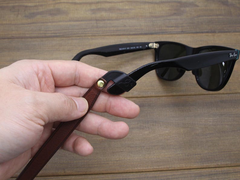 May include: Black sunglasses with a brown leather strap attached. The strap is held by a hand. The sunglasses are on a wooden surface. The strap has a gold-colored rivet.
