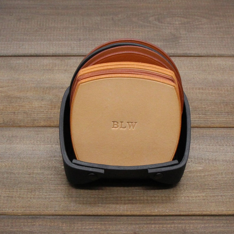 Puede incluir: Un juego de seis posavasos de cuero en un soporte negro. Los posavasos est&aacute;n apilados y el posavasos superior tiene las iniciales "BLW" grabadas.