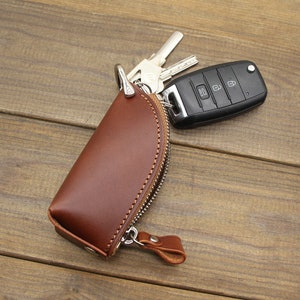 May include: A brown leather key holder with a zipper closure and a silver metal hook. The key holder is lying on a wooden surface with a car key attached to the hook.