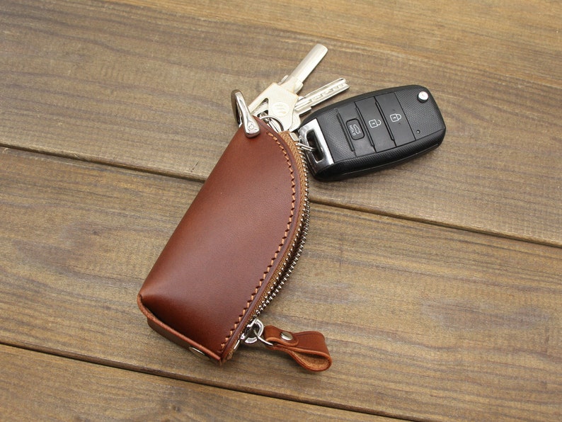 May include: A brown leather key holder with a zipper closure and a silver metal hook. The key holder is lying on a wooden surface with a car key attached to the hook.
