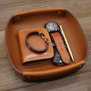 May include: A brown leather tray with a watch, a wallet, a pen, and a key. The tray has a stitched edge and is made of high-quality leather.