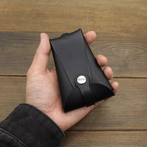 May include: A black leather wallet with a silver button closure. The wallet is folded in half and held in a hand.