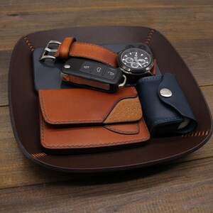 Puede incluir: Una bandeja de cuero marr&oacute;n con un reloj, una llave, una cartera y una peque&ntilde;a bolsa azul. La bandeja est&aacute; hecha de cuero y tiene un borde cosido.